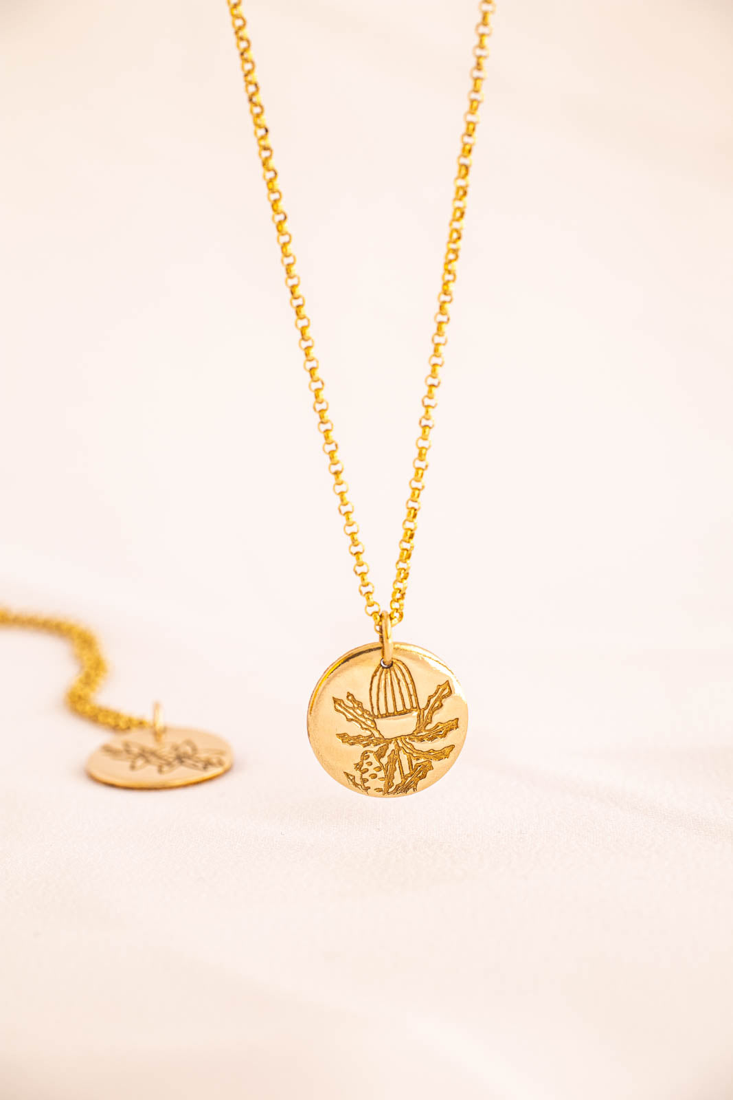 A gold-colored necklace featuring a round pendant with a floral design, specifically the Banksia flower, laid out on a plain surface.